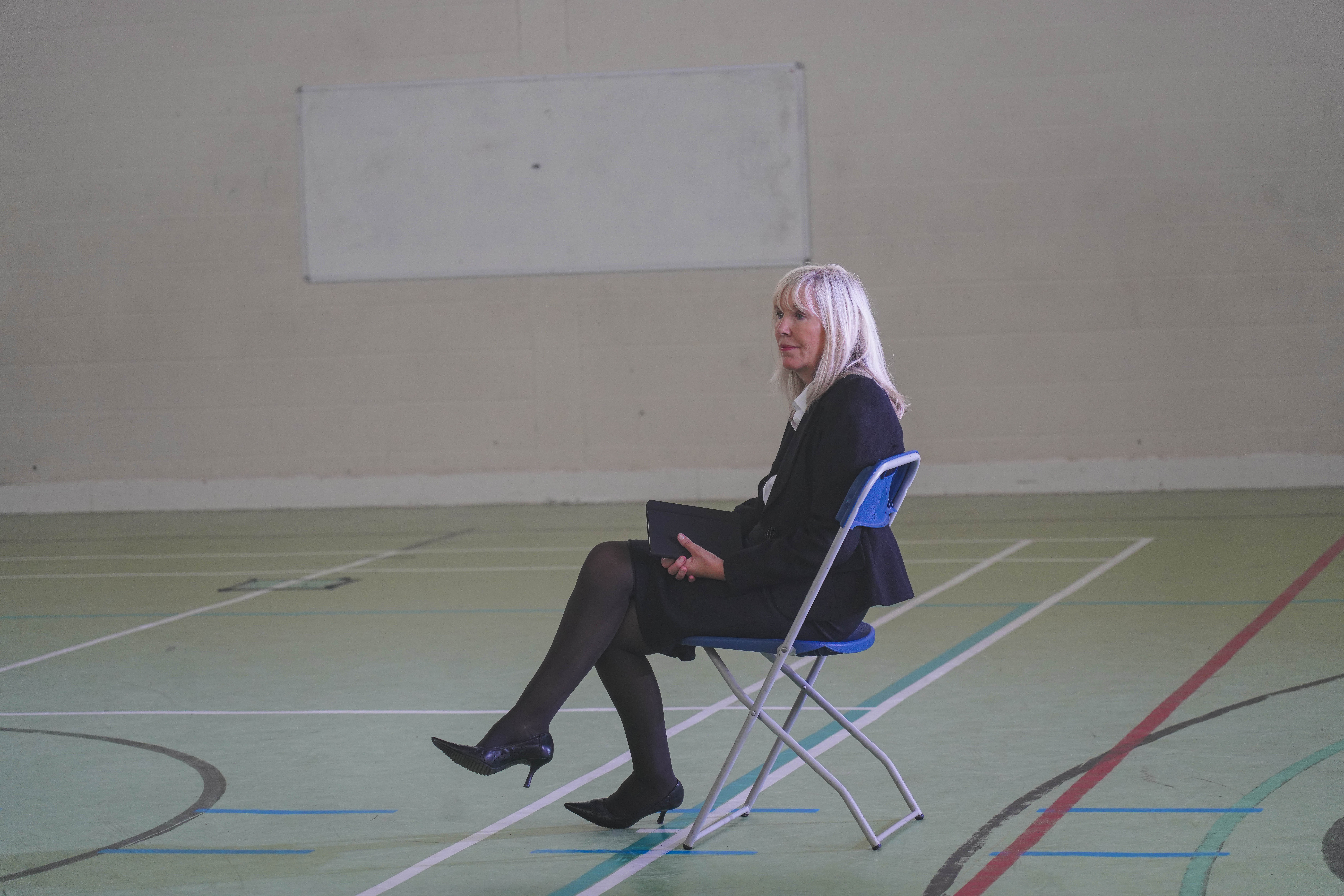Production still from Typical? showing a woman seated alone on a chair in a large empty sports hall.